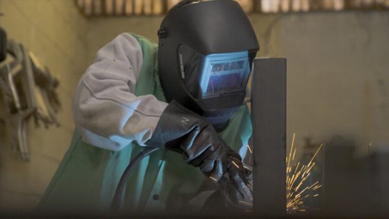 A person in a welding mask welding a piece of metal. 